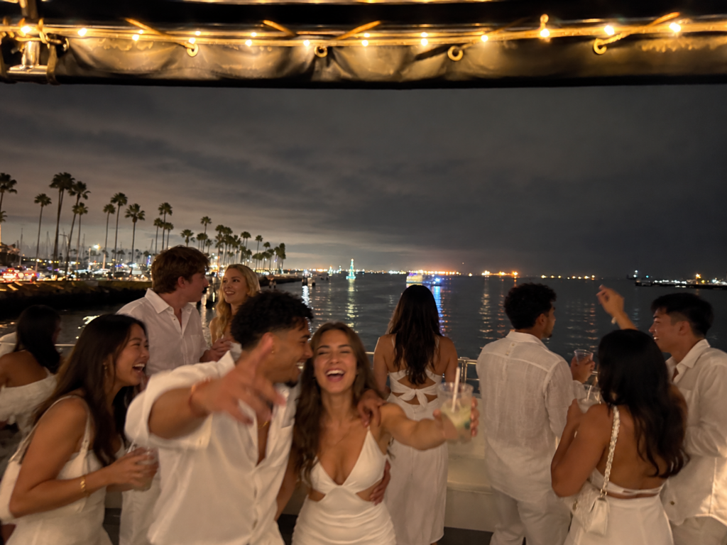 College students dressed in all-white outfits celebrating on a private yacht at night, with string lights and illuminated harbor views.